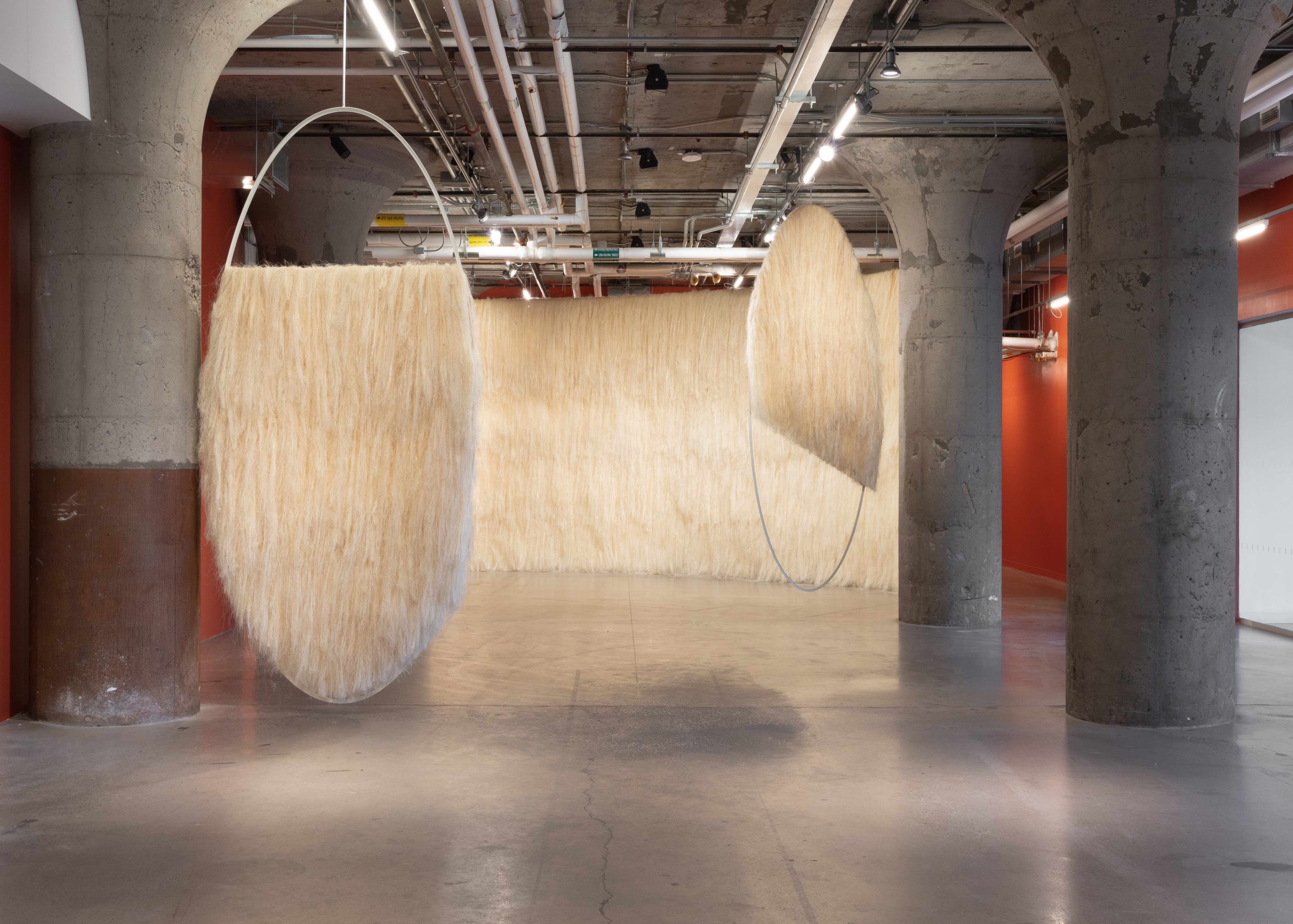 The large-scale installation Elliptical Field by Kipwani Kiwanga including two metal circles hanging from the ceiling, adorned with cream-coloured sisal fibre. The circles are set against a larger sisal fibre background in the same hue.