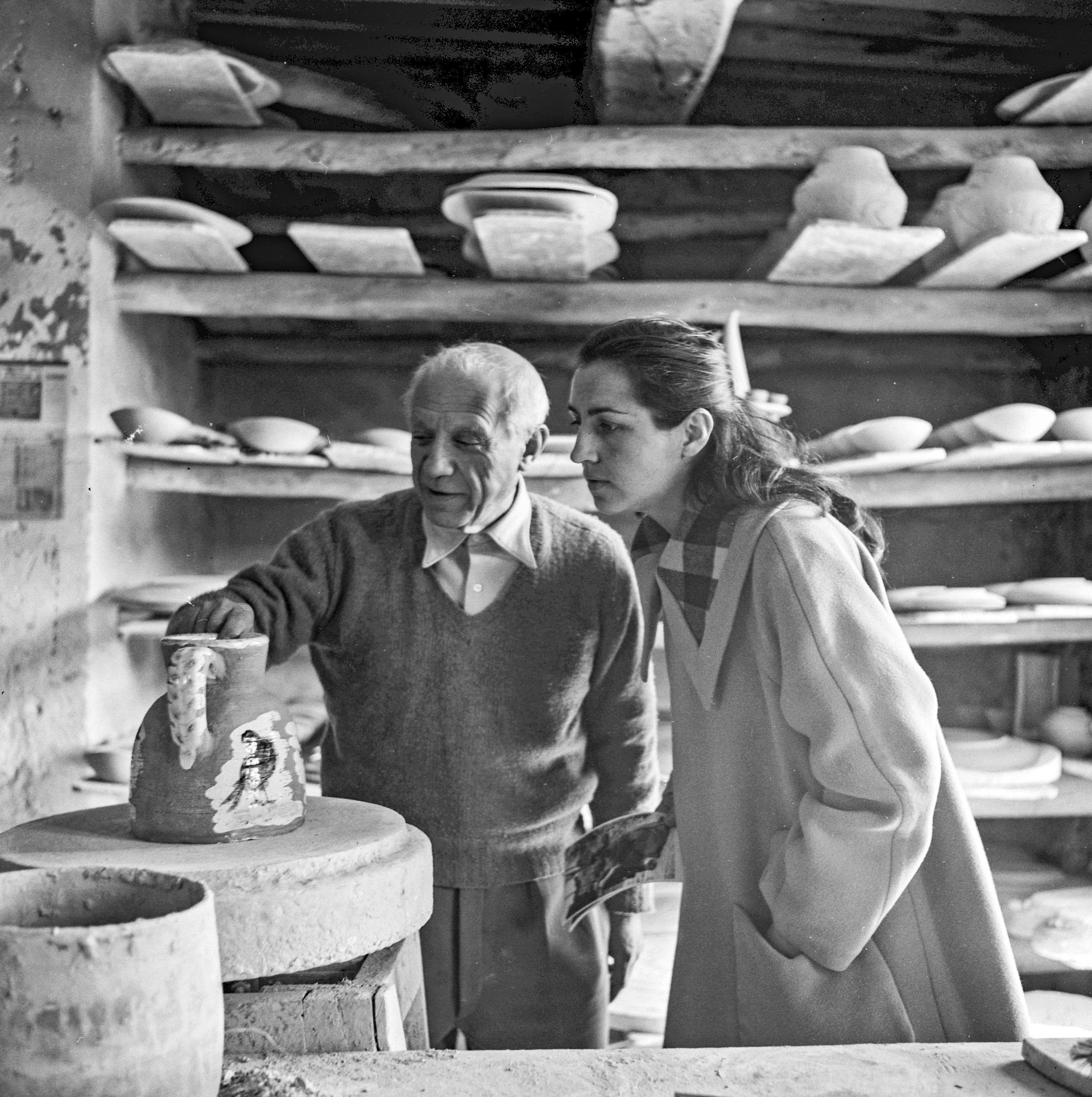 A black-and-white photo shots artist Pablo Picasso looking at a piece of pottery with Françoise Gilot at the Madoura Pottery Studio.