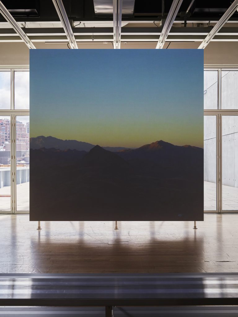 A square screen is set against a wall of windows in a gallery space with a wood floor. The screen depicts a lightly hilled landscape bathed in twilight.