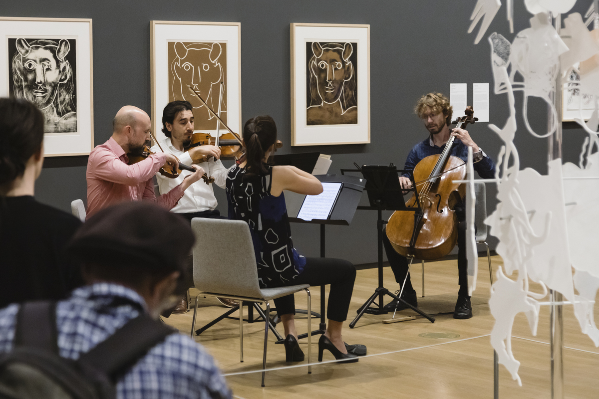 A group of classical musicians play music in front of picasso linocuts in an art gallery space
