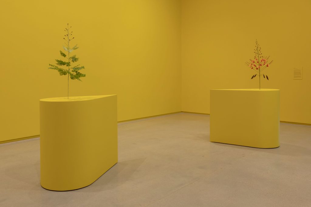 Two yellow teardrop-shaped plinths display paper flowers. The wall is also painted yellow behind them.