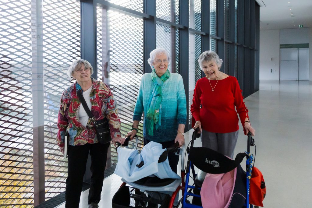 Three seniors walk together down a hallway in Remai Modern. Two use mobility aids.