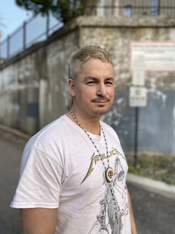 Wally Dion standing in front of a weathered stone wall, wearing a white Metallica t-shirt and a necklace with a circular pendant and feather. He has short, light-colored hair and a slight smile, with a relaxed and confident demeanor. The background features an urban setting with a fence and signage partially visible