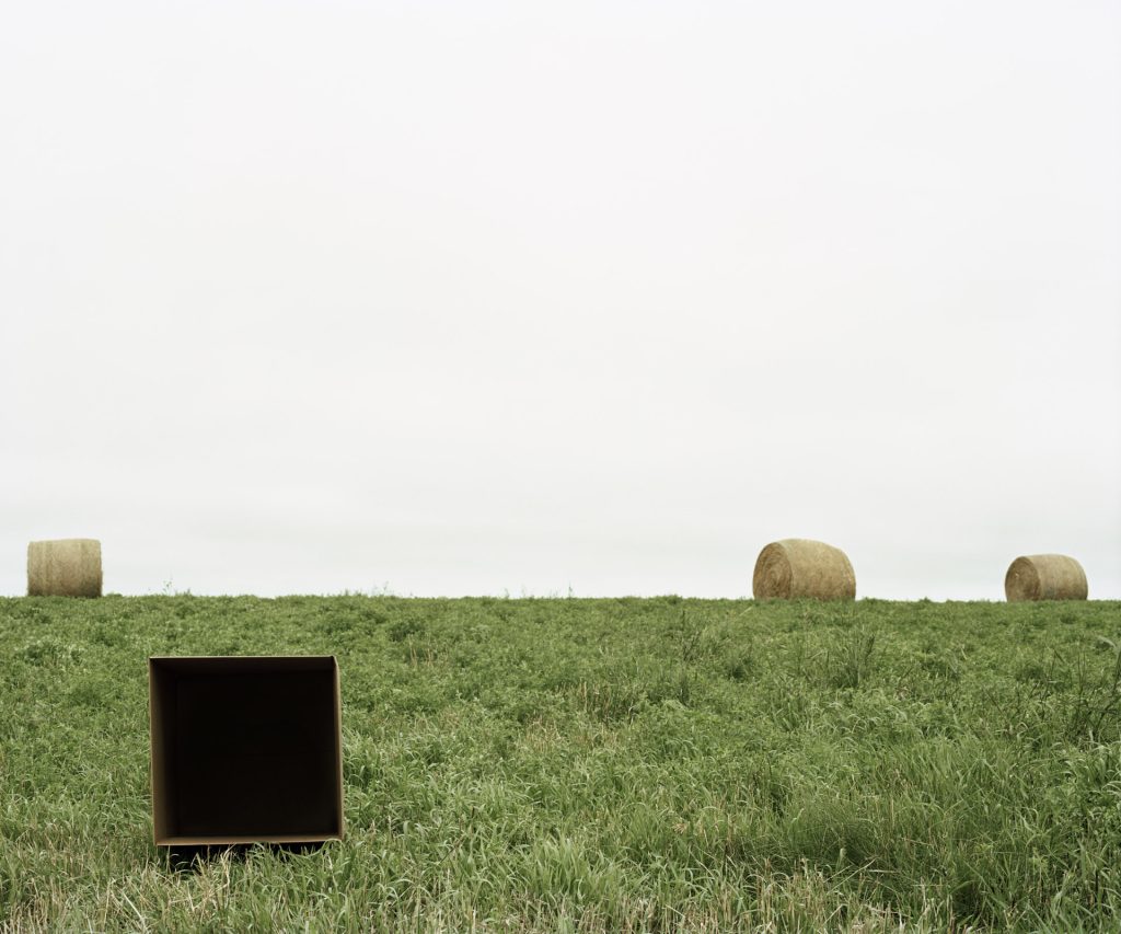 A landscape photograph of a grassy field under an overcast sky, with large round hay bales scattered in the distance. In the foreground sits an empty, dark, cubic box, contrasting with the natural surroundings.