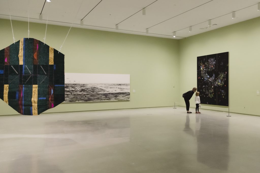 A visitor and a child lean in to look closely at a large artwork in a spacious gallery with pale green walls and several large-scale works on display.