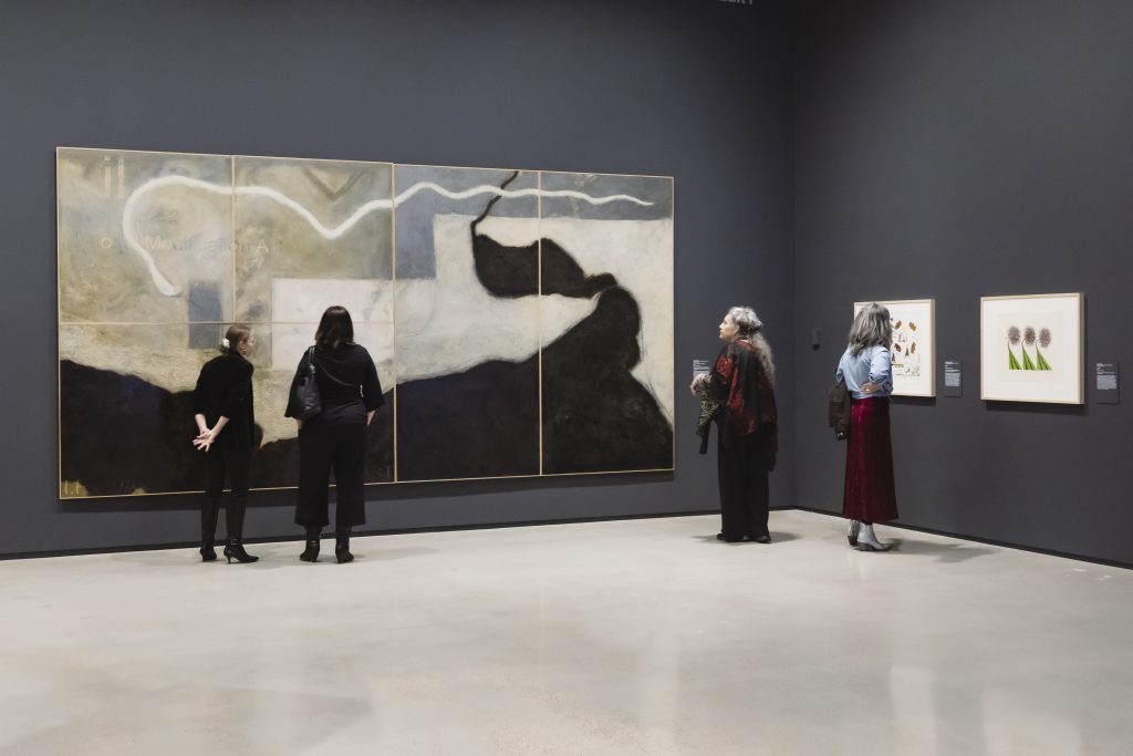 Visitors stand in a gallery with dark blue walls, viewing a large multi-panel painting and framed works hung nearby.