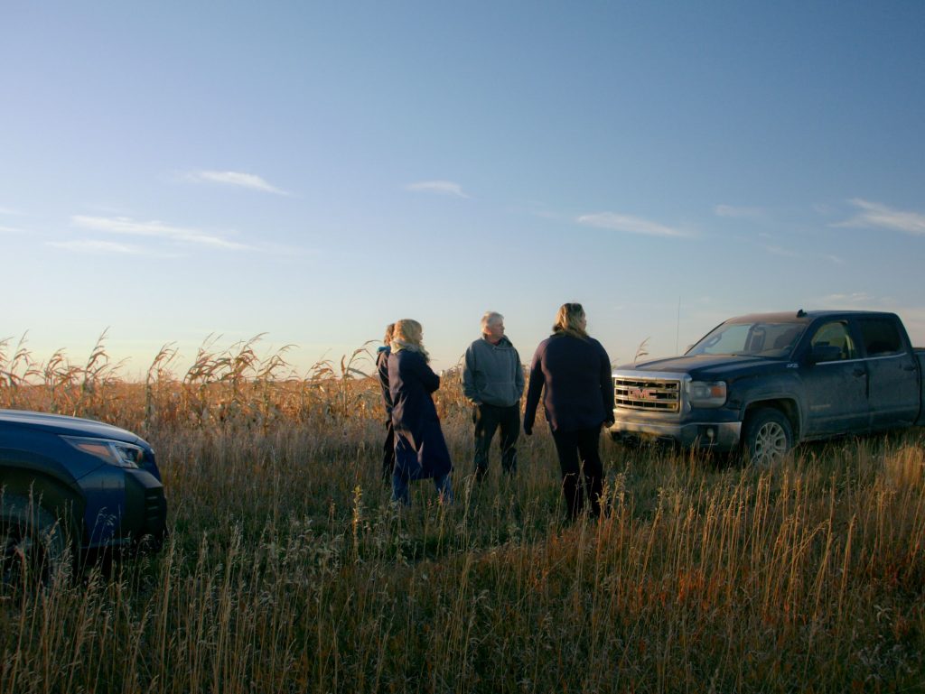 Three people stand in a grassy field between two parked vehicles, a blue SUV and a dusty grey pickup truck, under a clear sky at sunset. They appear to be in conversation, surrounded by tall dry grass and distant corn stalks.