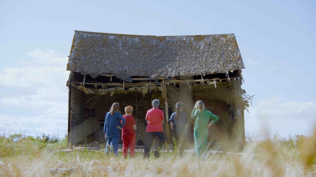 Five people stand in a grassy field facing a large, weathered barn with a partially collapsed roof and exposed beams. They wear brightly colored clothing—red, blue, and green—contrasting with the muted tones of the deteriorating wooden structure under a clear blue sky.