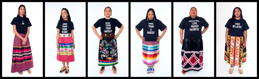 Six indigenous women stand in front of white backgrounds in a
horizontal row. each wears a colourful skirt, some wear moccasins, each also wears a
black t shirt with white text. In order from left to right they read: I AM, MORE THAN YOUR
DESIRE, MORE THAN YOUR FANTASY, MORE THAN A MASCOT, ANCESTRAL LOVE
PRAYER SACRIFICE, YOUR RELATIVE