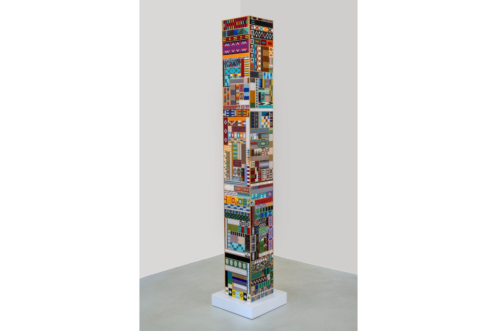 A large columnar sculpture made up of a patchwork of beaded panels. Each is sewn into different geometric patterns, many are highly reflective