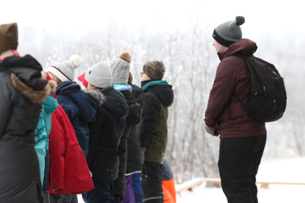 A group of people dressed in winter coats and knit hats stand outdoors on a snowy day, looking out toward a frosty landscape, while a person with a backpack stands slightly apart to the right.
