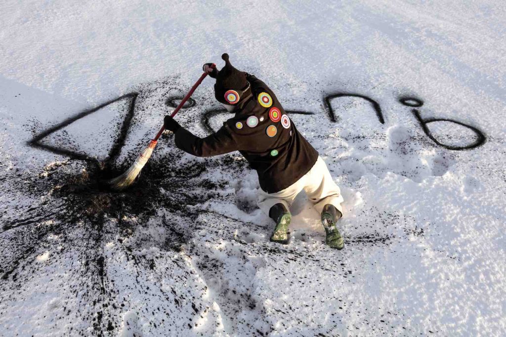 A photo work by artist Meryl McMaster shows a figure kneeling in the snow and writing in syllabics using dirt.