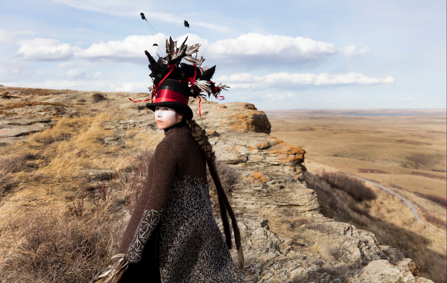 A photograph by Meryl McMaster features a rocky, grassland landscape and a self portrait of the artist wearing a black cape, feathered top hat and a band of white makeup across her eyes.