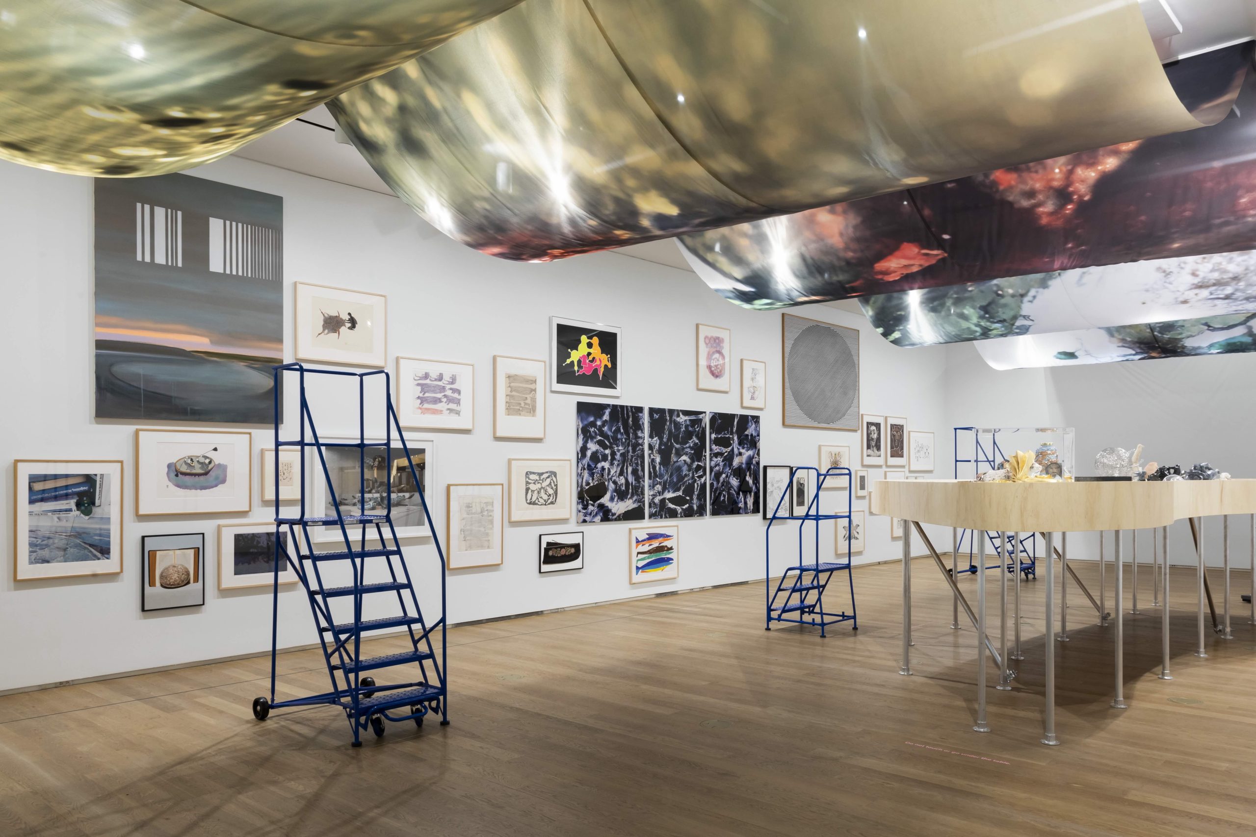 A contemporary art gallery installation with a variety of framed artworks displayed on a white wall, arranged in a salon-style format. The artworks include abstract and representational pieces in different mediums. Blue metal step ladders are positioned in the space, and overhead, large translucent fabric panels with abstract designs are suspended from the ceiling. A sculptural table on silver legs displays a collection of objects and materials. The room features wooden flooring and soft ambient lighting.