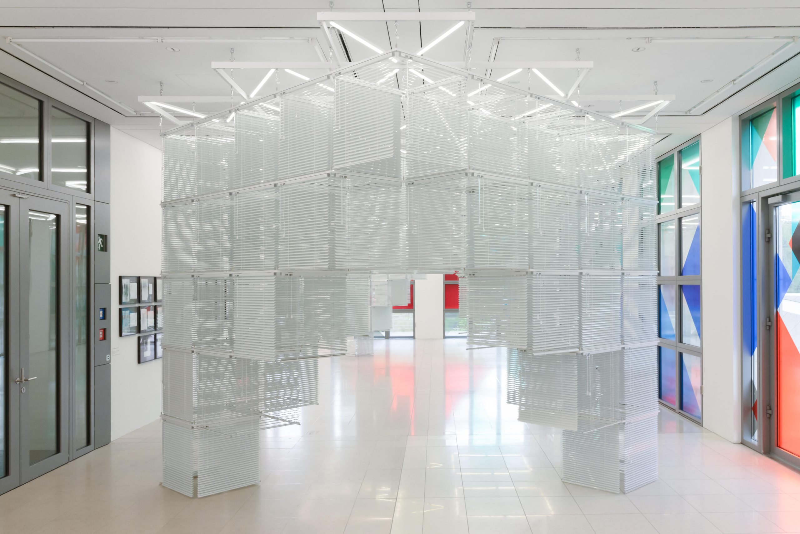 A structure made from white venetian blinds hangs from the ceiling to the floor in a gallery space.