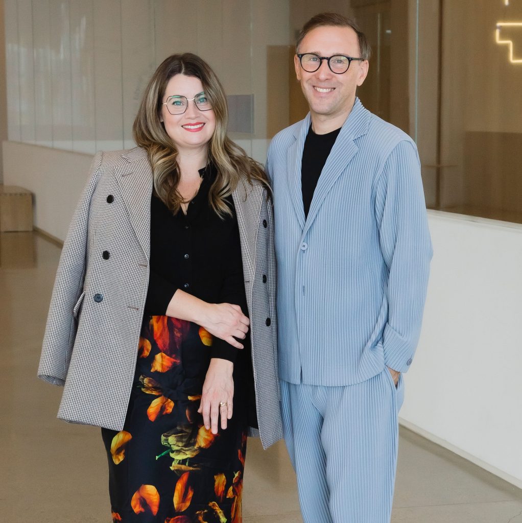 Two people stand side by side indoors in a modern, light-filled gallery space. Both are smiling at the camera. One person wears a patterned blazer over a black top and a floral skirt, and the other wears a light blue textured suit with glasses.