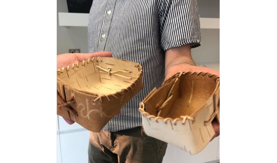 A person holding two small birch bark baskets, one in each hand, showing the handcrafted details and natural materials used in their construction. The baskets are stitched together with natural fibers, and the background suggests an indoor setting.