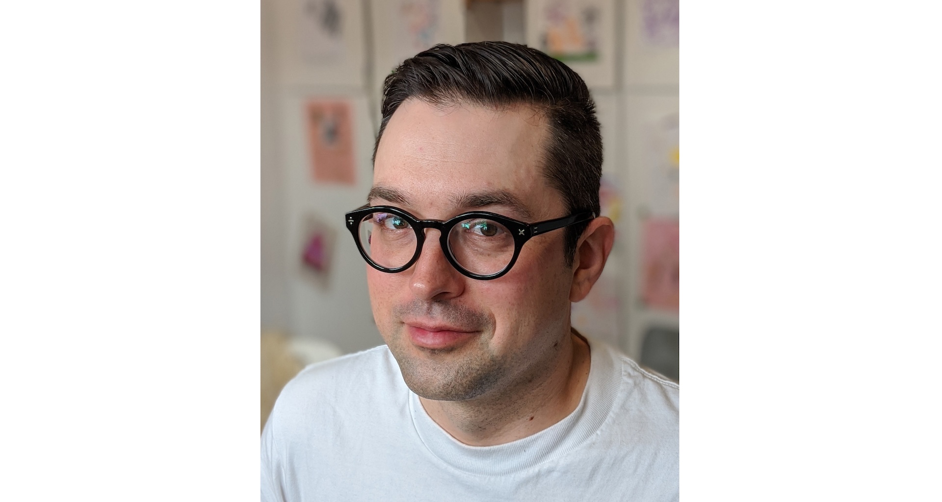 "Portrait of Jacob Semko, wearing black-rimmed glasses and a white shirt, standing in front of a wall decorated with various drawings."