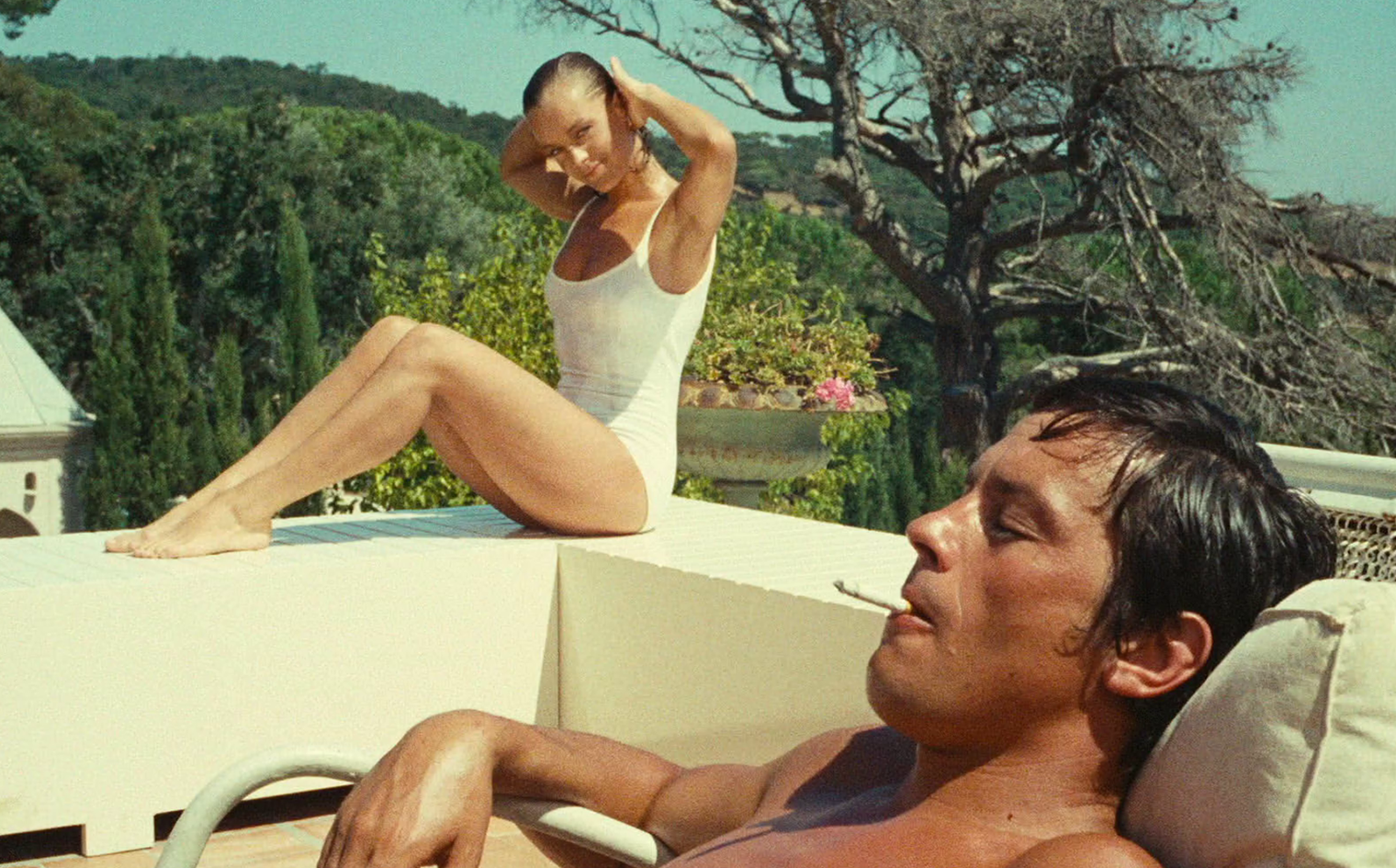 Two people relaxing by a poolside in a sunny, outdoor setting. A woman in a white swimsuit is seated on the edge of the pool, adjusting her hair and looking towards the camera with a relaxed expression. She is positioned higher than the man, who is lying on a pool chair in the foreground. The man is shirtless, with dark hair, and is smoking a cigarette while leaning back comfortably with his eyes closed. The background features lush greenery and a clear sky, evoking a serene and leisurely atmosphere.