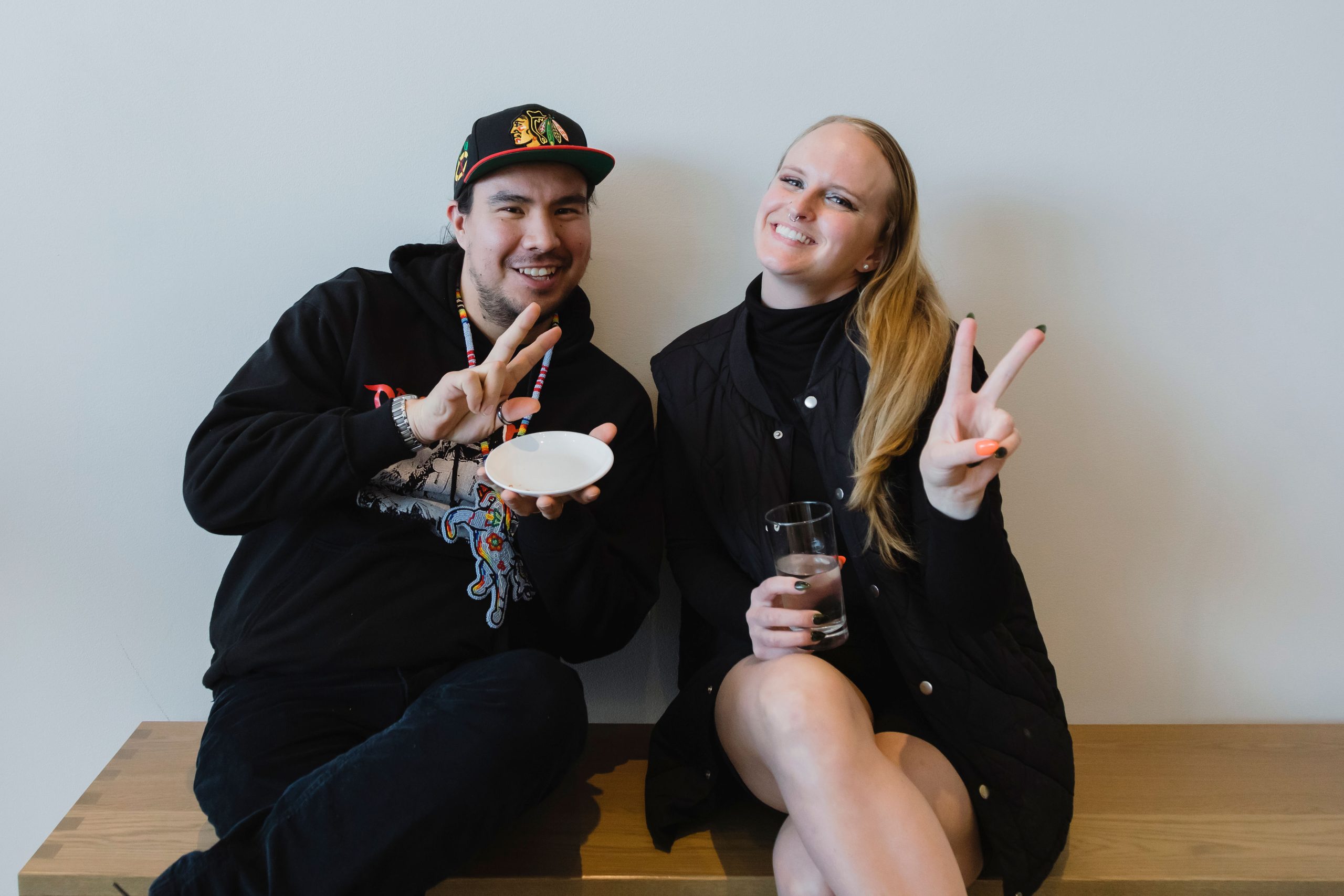 Two people are seated on a wooden bench, smiling and flashing peace signs at the camera. The person on the left is wearing a black hoodie, a colorful beaded necklace, and a cap with a sports logo. The person on the right is wearing a black jacket and holding a glass of water.