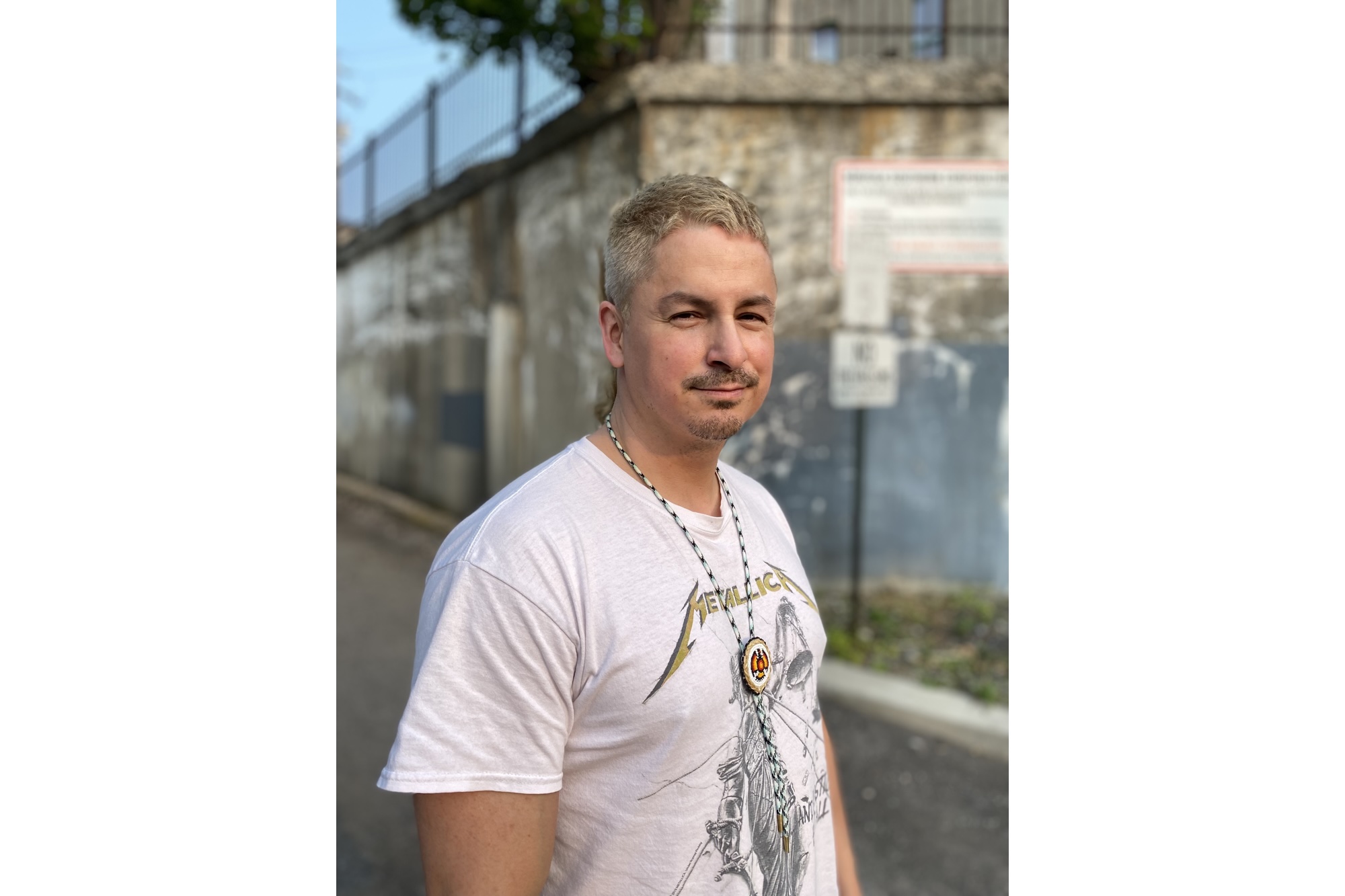 Wally Dion standing in front of a weathered stone wall, wearing a white Metallica t-shirt and a necklace with a circular pendant and feather. He has short, light-colored hair and a slight smile, with a relaxed and confident demeanor. The background features an urban setting with a fence and signage partially visible