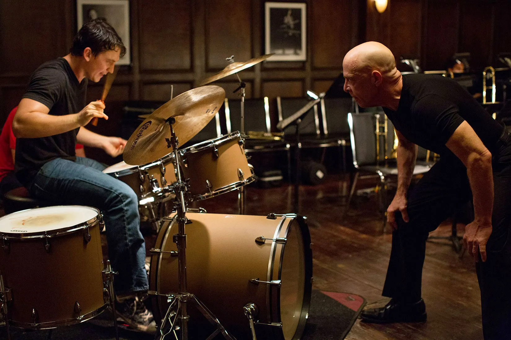 "A man intensely playing drums with an instructor in a music rehearsal room."