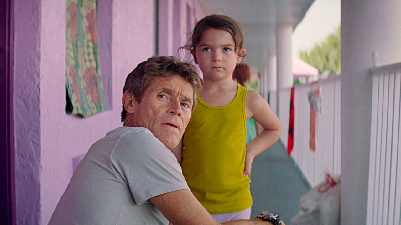 A man with a concerned expression sits in a chair, turning slightly to look behind him. A young girl in a yellow tank top stands next to him with a serious expression. They are in a pastel-colored corridor, likely of a motel or apartment building. The setting suggests a moment of tension or contemplation.