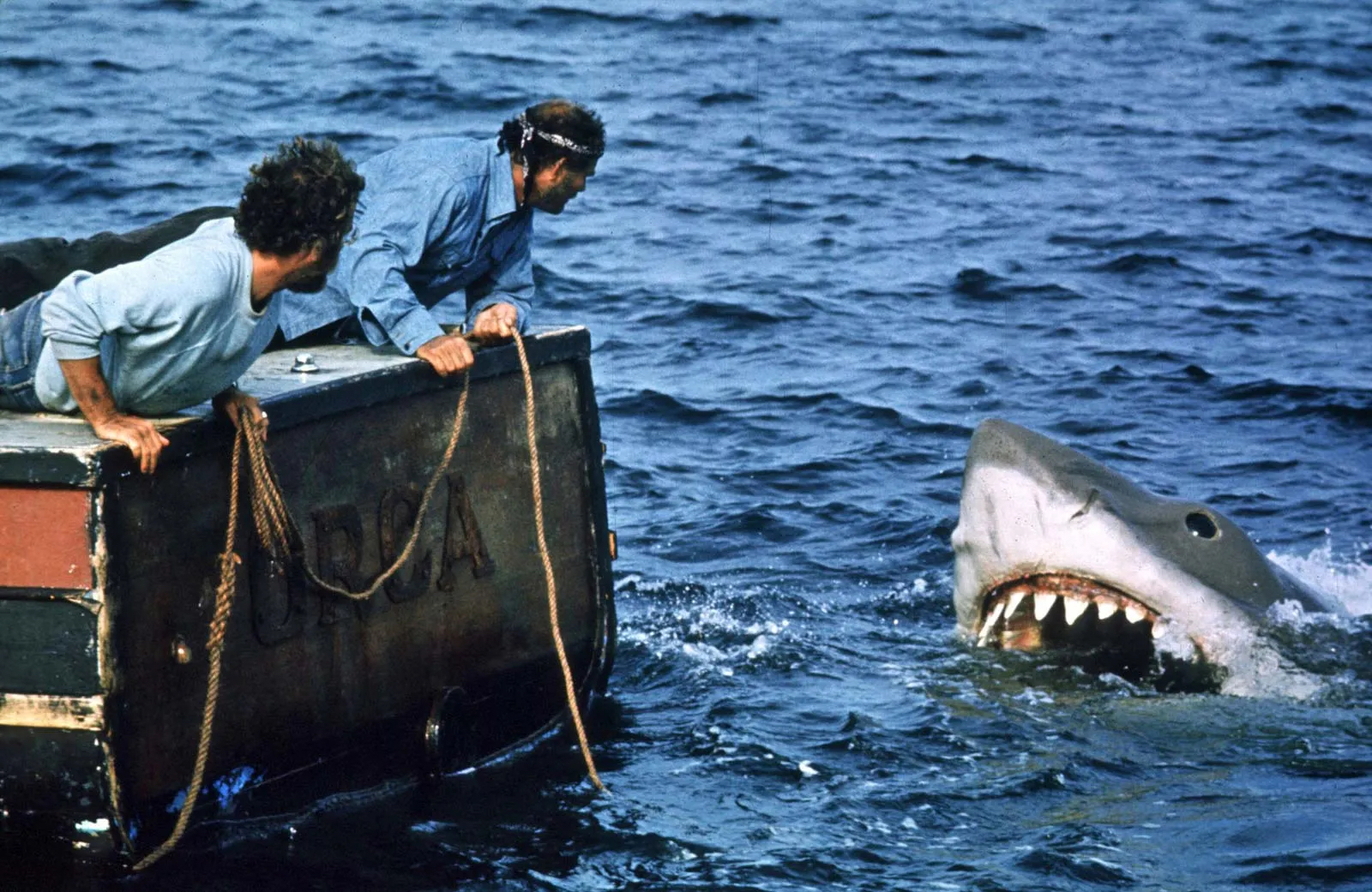 Two men are on a boat named “Orca,” leaning over the edge and staring at a large, menacing shark that is emerging from the water. The shark has its mouth open, showing sharp teeth, and appears to be very close to the boat. One man is wearing a light blue shirt, and the other is wearing a dark blue shirt with a bandana on his head. The scene takes place in the open ocean, with waves visible in the background, creating a sense of tension and danger.