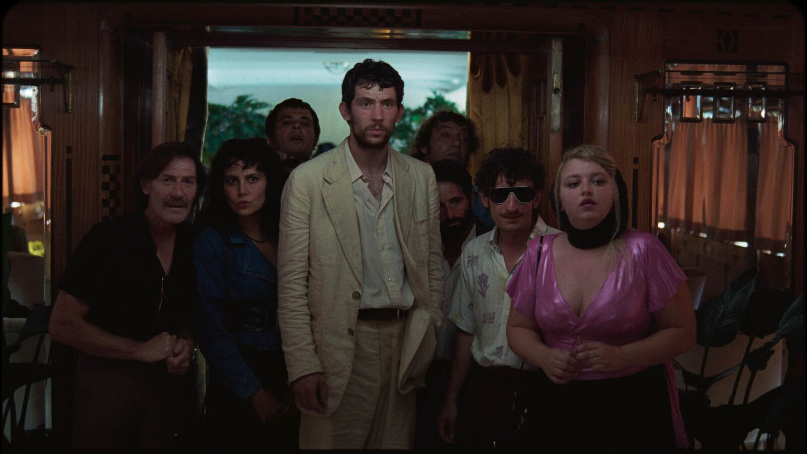 A group of people stands closely together in a dimly lit room, looking intently towards something off-camera. The central figure, a man in a light-colored suit with a disheveled appearance, stands out prominently. To his right, a woman in a bright pink blouse with a low neckline and a black headscarf looks towards the camera with a concerned expression. Another man with dark hair and sunglasses stands next to her, wearing a light-colored shirt. The group includes both men and women, all displaying a mix of curiosity and apprehension. The background features wooden paneling and curtains, suggesting an interior setting.