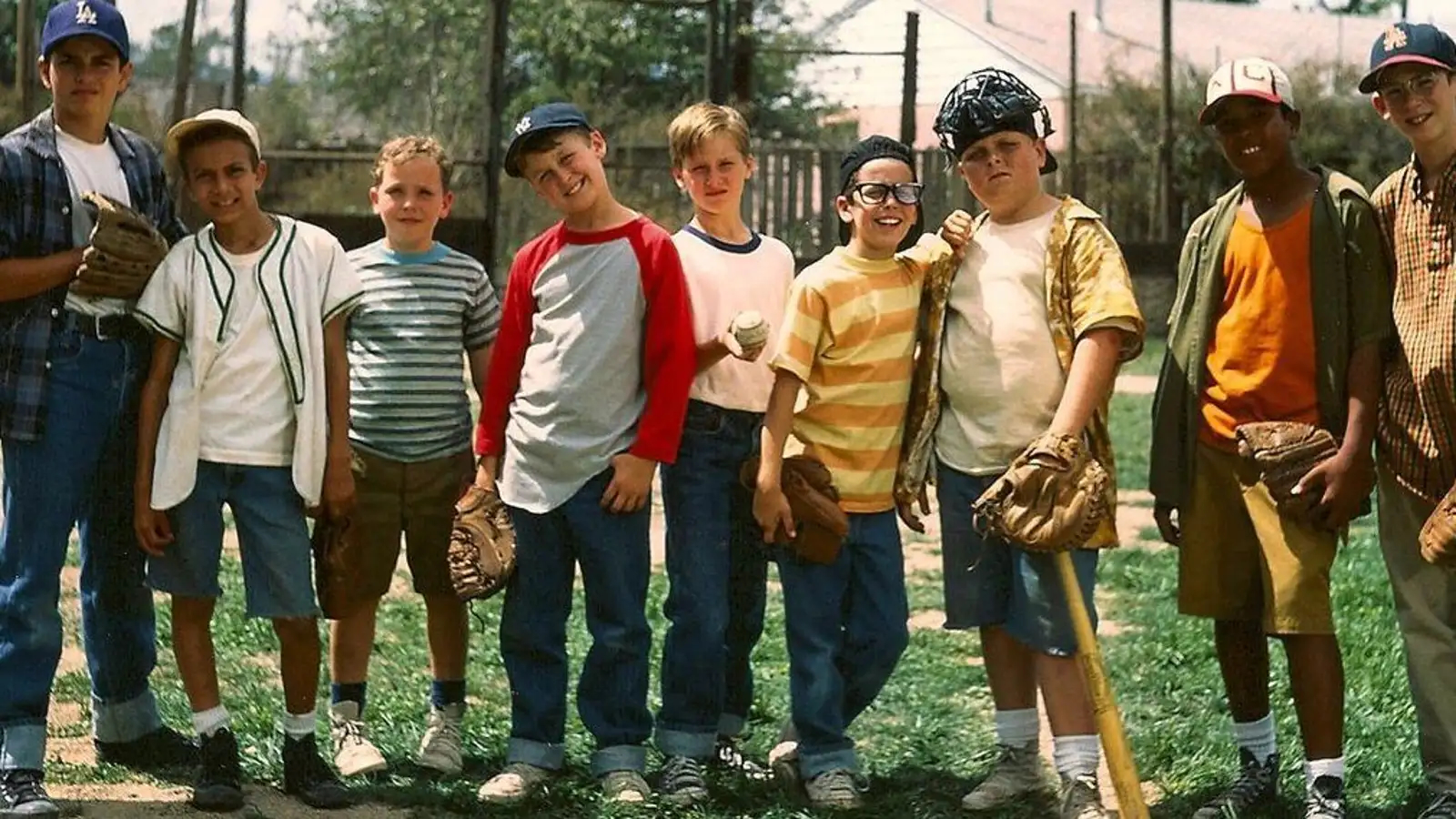 A group of young boys stands together on a grassy field, posing for a photo in their casual baseball attire. Each boy is holding a piece of baseball equipment, such as gloves, a bat, or a ball. They are dressed in various shirts and caps, reflecting a mix of styles from the era. The boys are smiling and standing close to each other, showing camaraderie and the carefree spirit of childhood. The background includes a fence and some trees, indicating they are in a neighborhood or park setting. The image captures a nostalgic, playful moment from a classic American pastime.