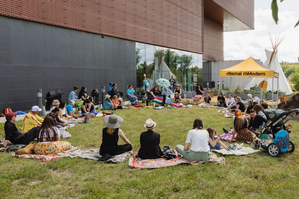 A group of people sit outside Remai Modern in a circle.