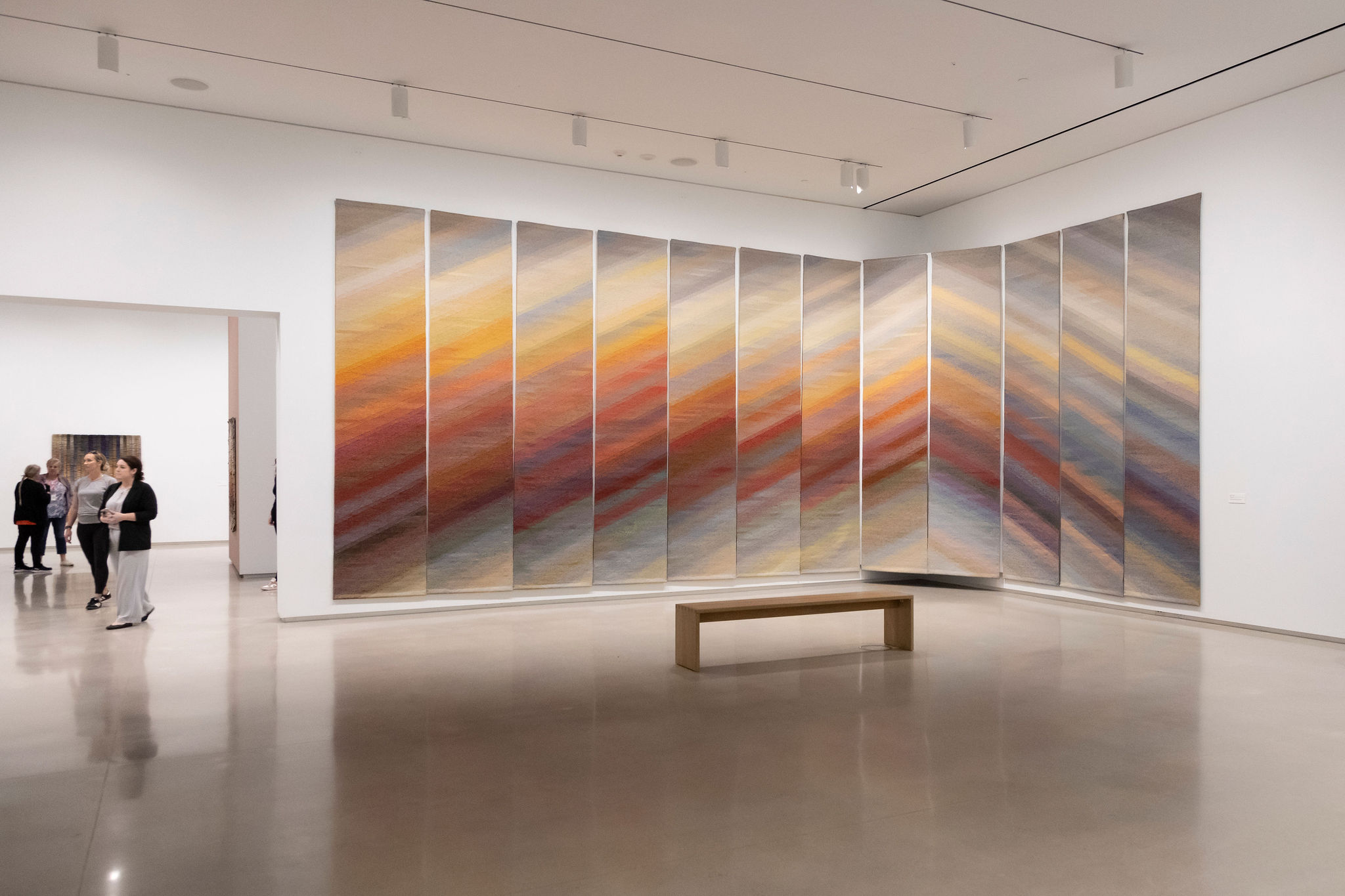 Museum visitors walk through a contemporary gallery space featuring a large-scale, multi-panel artwork with soft, gradient hues of orange, red, blue, and grey. The installation, displayed on a curved wall, creates a sweeping visual effect. A minimalist wooden bench is positioned in front of the artwork, inviting viewers to sit and engage with the piece.