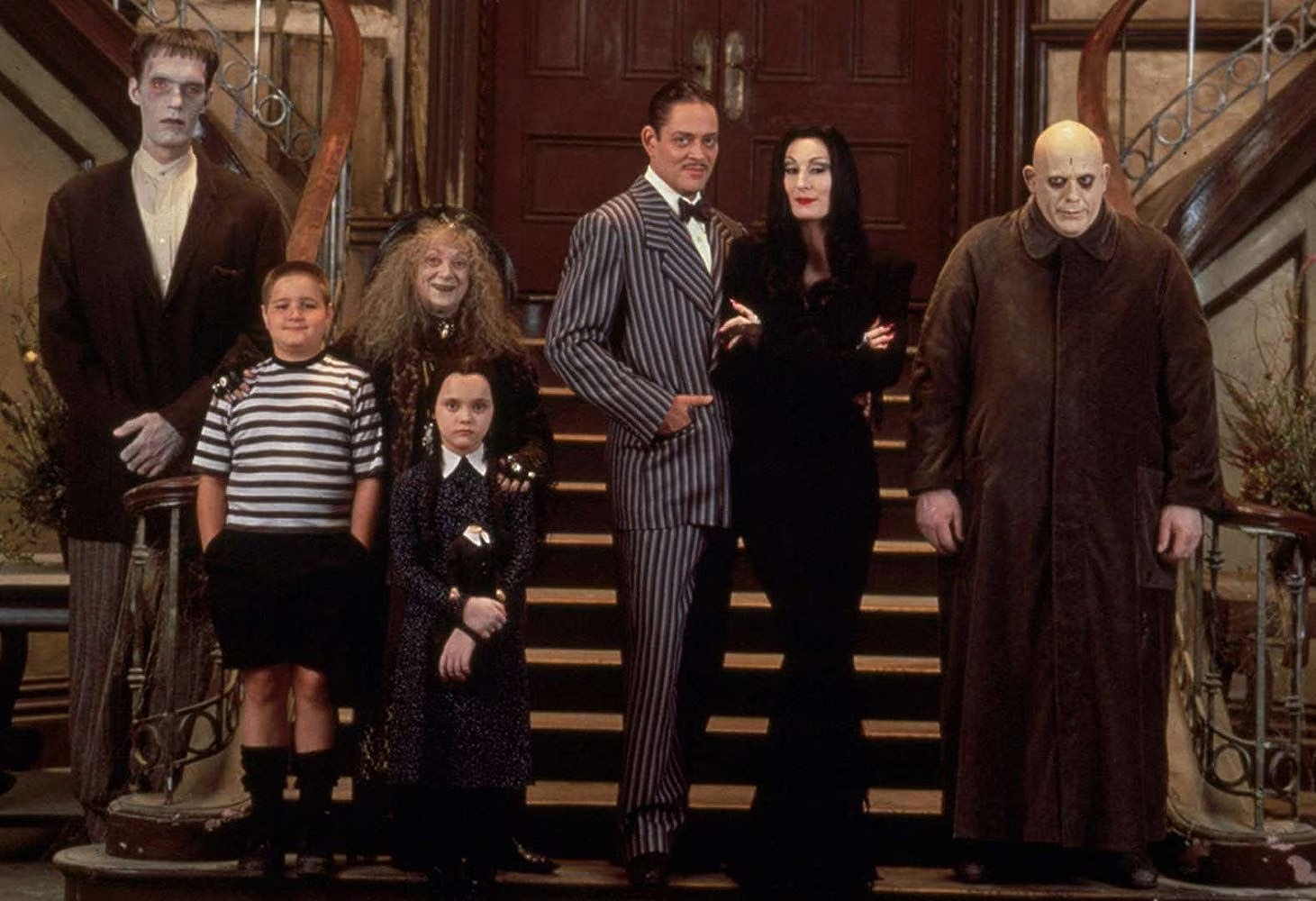 A group photo of the iconic characters from The Addams Family (1991) film, featuring Lurch, Pugsley, Wednesday, Grandmama, Gomez, Morticia, and Uncle Fester, all standing in front of the grand staircase in their gothic mansion. The characters are dressed in their signature quirky and macabre outfits, with serious and stoic expressions that embody their darkly comedic personalities.