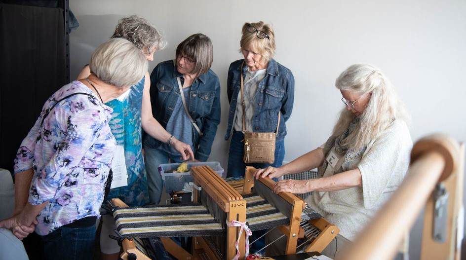 People gather around a traditional weaving loom, observing a demonstration. The woman at the loom, with long white hair and glasses, is focused on her work, guiding threads through the loom. The other women, dressed casually in denim and light clothing, watch attentively, some leaning in to get a closer look.