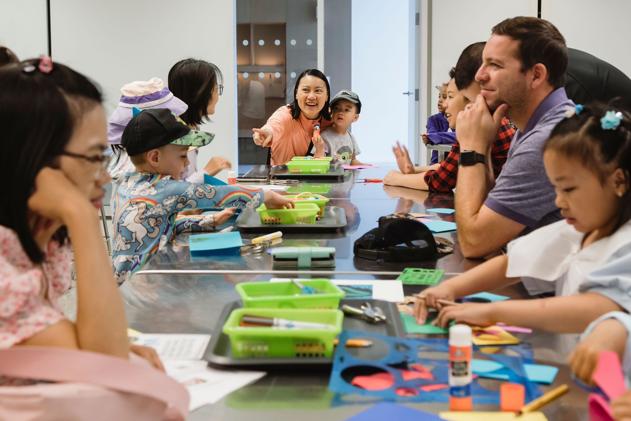 A group of adults and children gather around a table in a brightly lit room for an arts and crafts activity. Children are focused on creating with colorful paper, glue, and markers, while an adult in the center engages with the group, smiling and pointing. The scene captures a lively and creative atmosphere with participants of various ages.