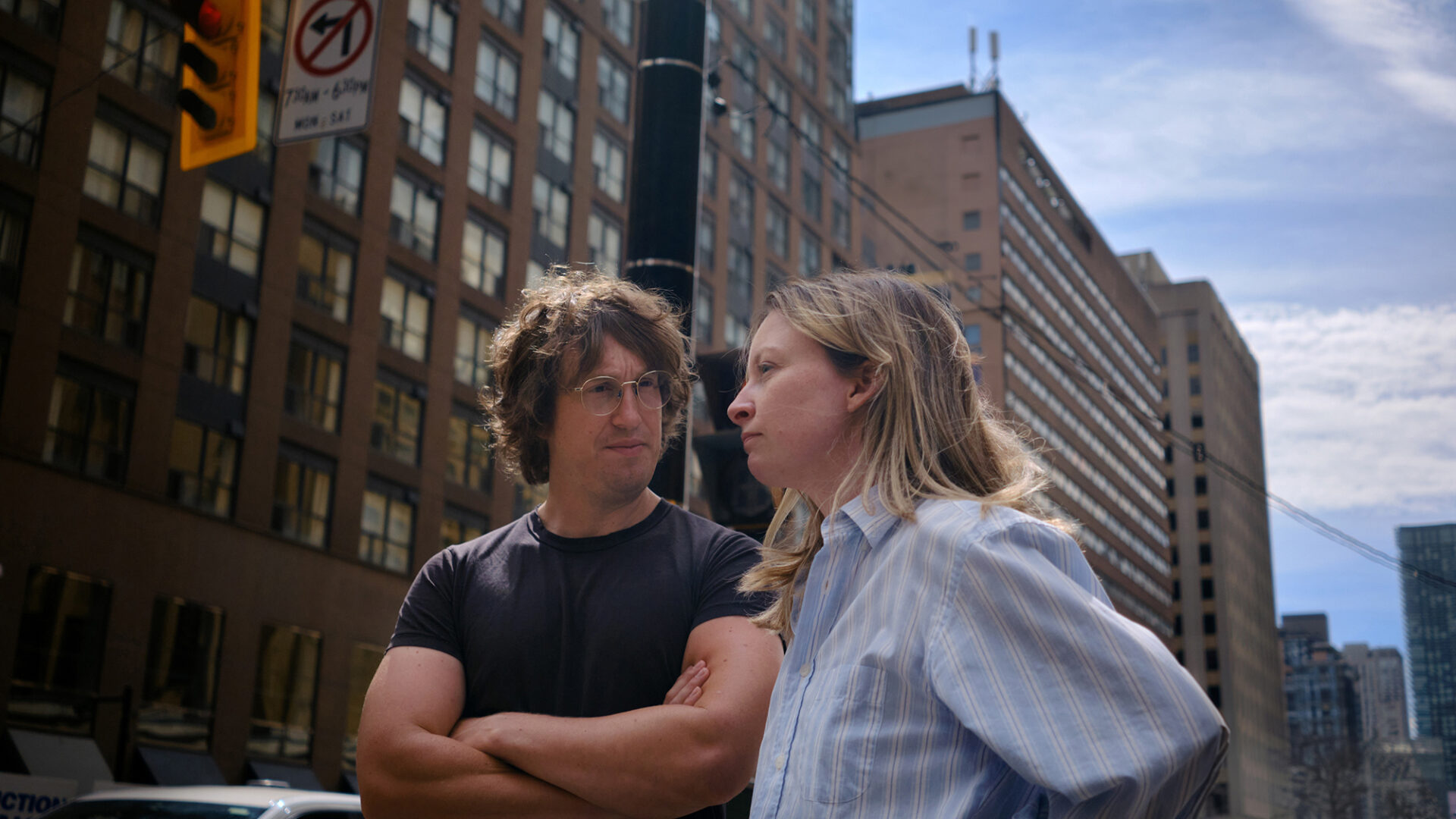 A man and a woman stand on a city street in conversation, surrounded by tall buildings under a bright, partly cloudy sky. The man, wearing glasses and a black t-shirt with crossed arms, looks intently at the woman, who is wearing a striped button-down shirt and appears to be speaking. A traffic sign and urban details in the background suggest a bustling metropolitan setting.