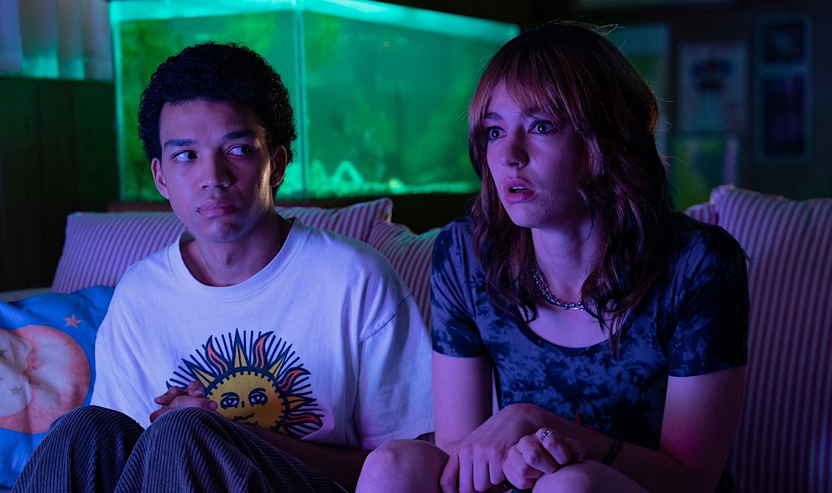 A man and woman sit on a couch, uncertain, in the glow of a TV, with a fish tank behind them.