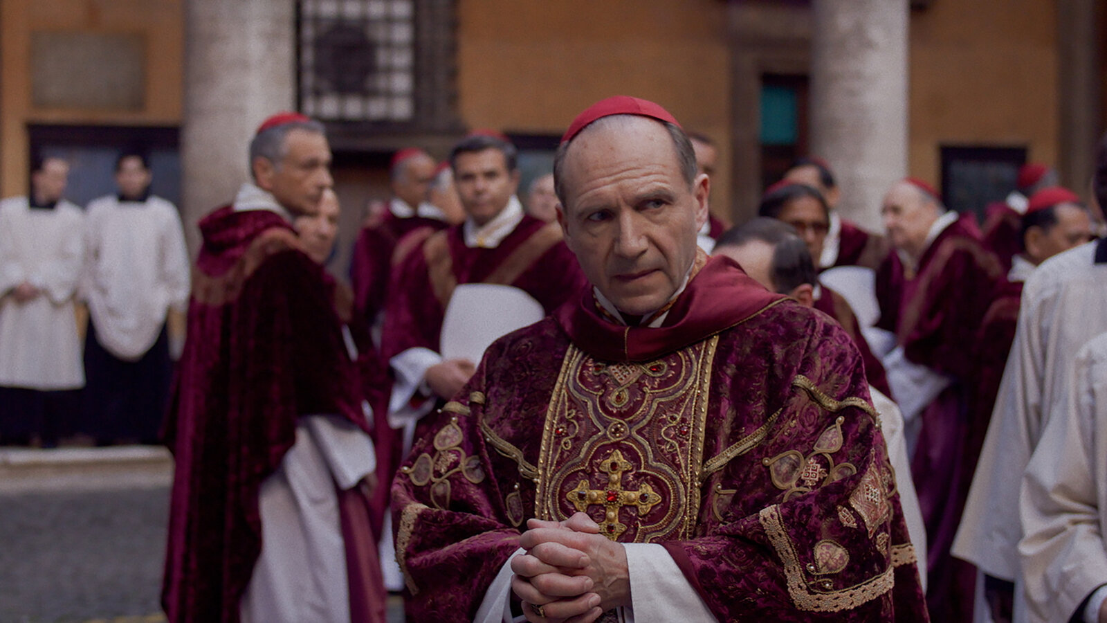 A solemn scene depicting a cardinal dressed in ornate red and gold vestments, surrounded by other clergy members in similar ceremonial robes, in an outdoor setting that suggests a historical or religious event. The cardinal appears to be deep in thought, with his hands clasped, as others mill about in the background.