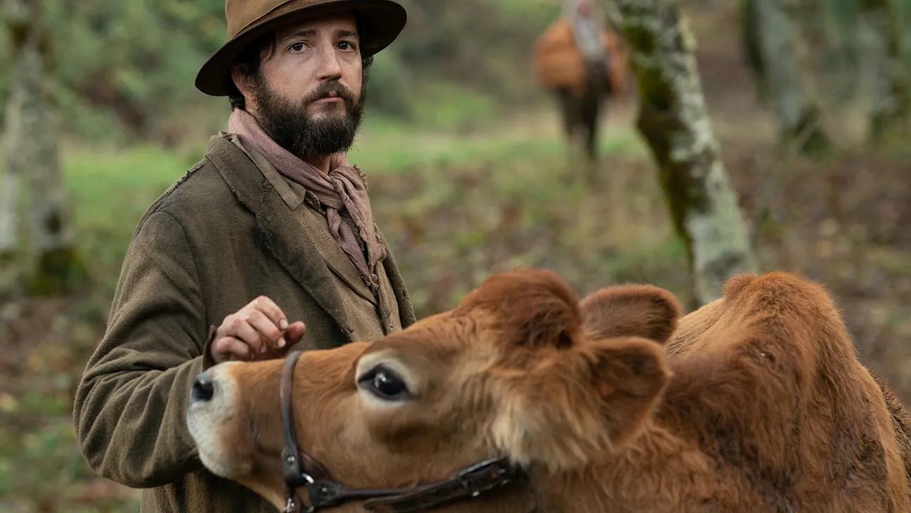 A bearded man in a hat stands beside a cow in a pastoral setting, showcasing a moment of rural life.