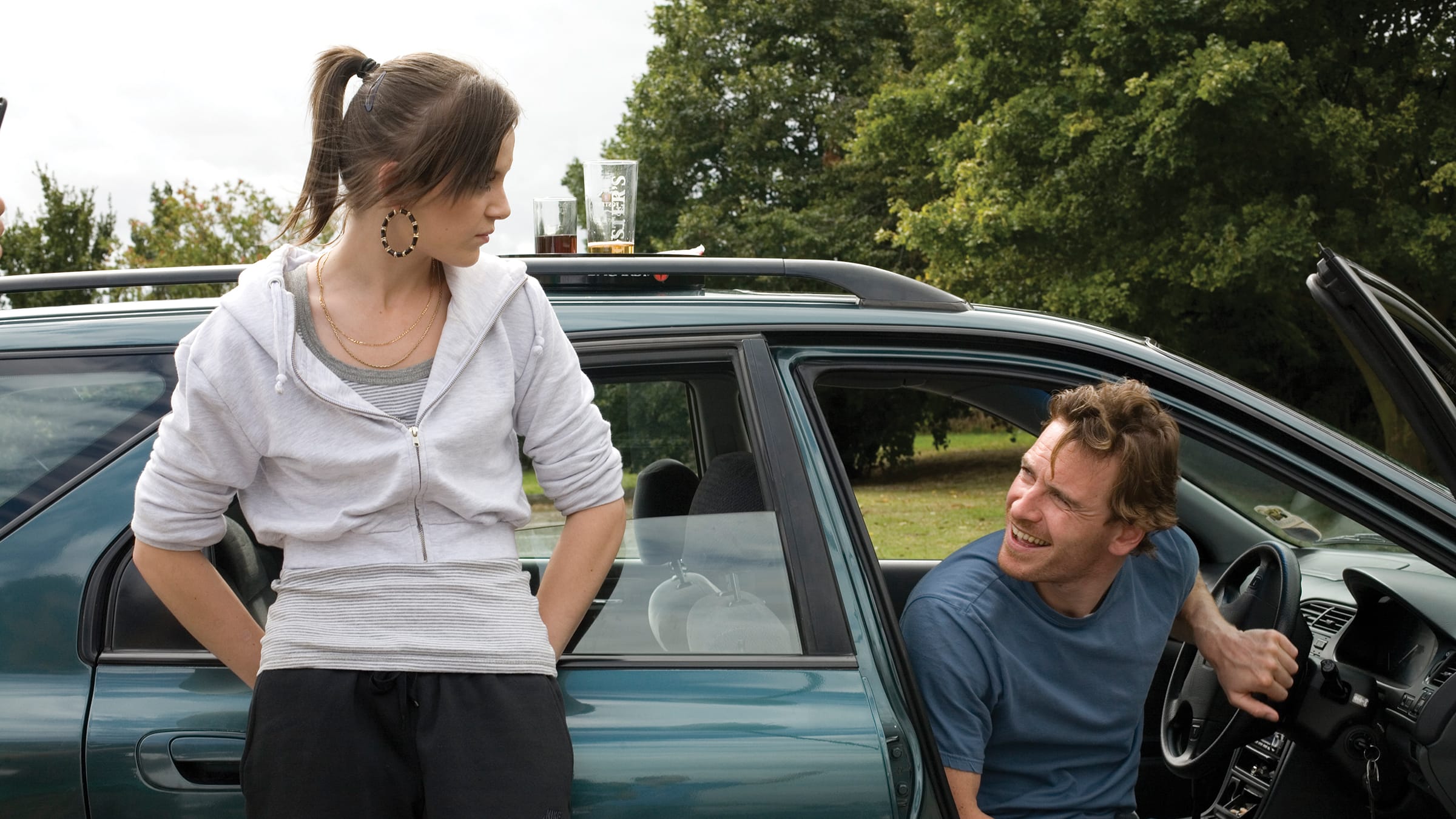 A man and woman stand beside a parked car, smiling and enjoying each other's company in a sunny outdoor setting.