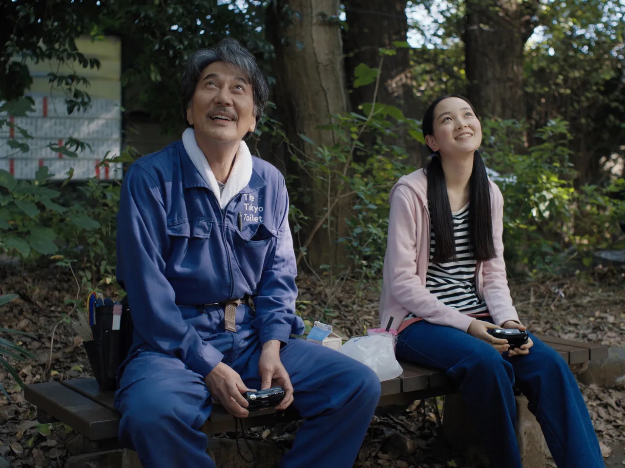 An outdoor scene featuring an older man and a young woman sitting on a wooden bench surrounded by greenery. The man, dressed in a blue work uniform with "The Tokyo Toilet" embroidered on it, holds a small device and gazes upward with a warm, thoughtful expression. The young woman, wearing a pink hoodie over a striped shirt and blue jeans, also looks upward with a gentle smile, holding a similar device. The serene forest setting, with sunlight filtering through the trees, adds a tranquil and reflective mood to the moment.