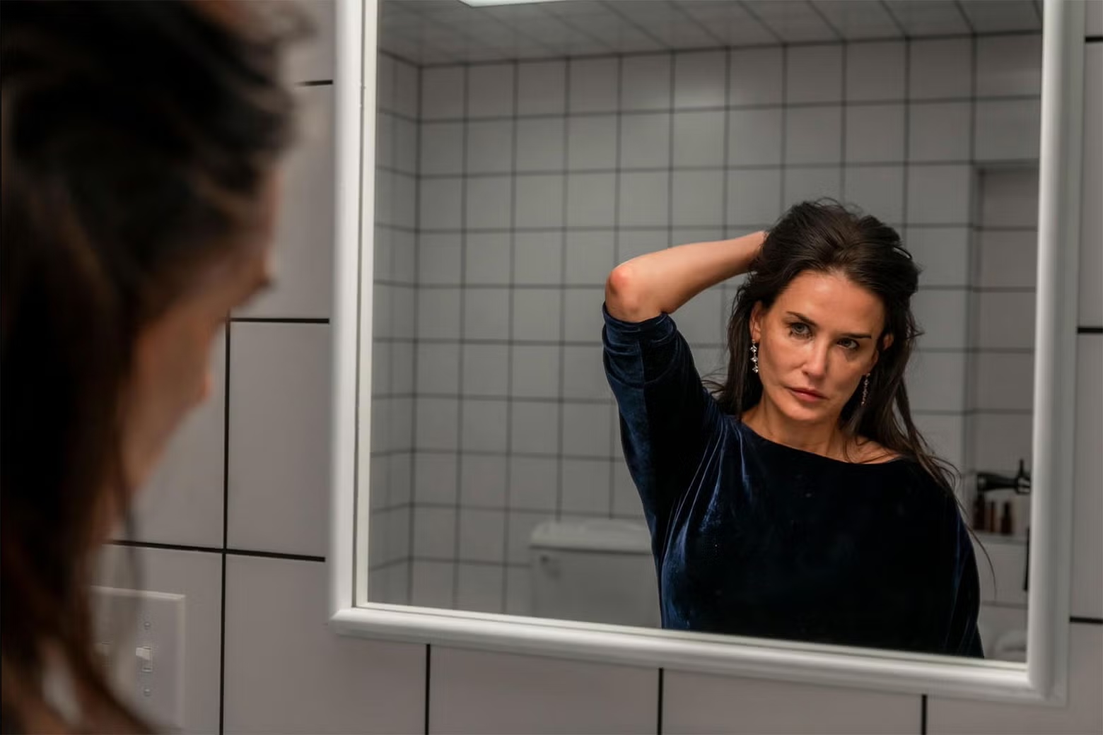 A woman with a contemplative expression looks at herself in a bathroom mirror. She is wearing a dark velvet dress and has her hand resting on the back of her head, adjusting her hair. The room features white tiled walls, and the mirror frames her reflection, emphasizing her introspective mood.