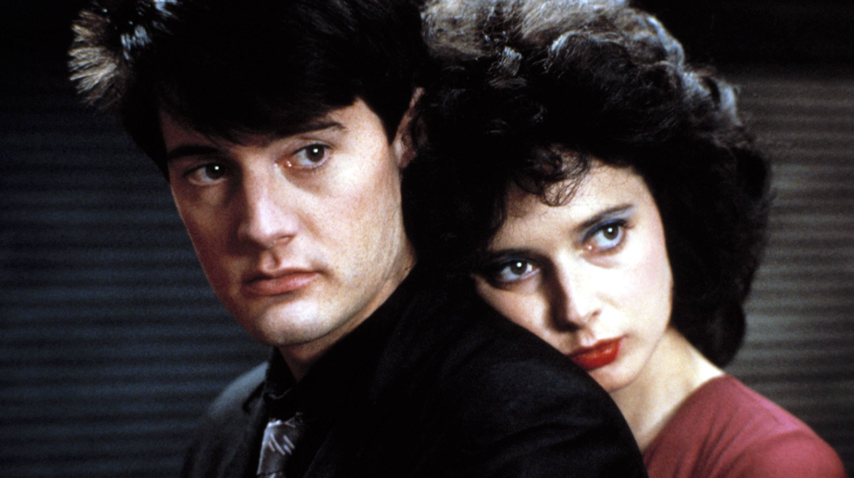 A close-up of a man and a woman in an intimate and dramatic moment. The man, with short dark hair, looks off to the side with a serious and slightly tense expression. The woman, with curly dark hair and striking red lipstick, rests her head on his shoulder, gazing directly forward with a pensive and intense look. The lighting emphasizes the shadows and adds a mysterious atmosphere.