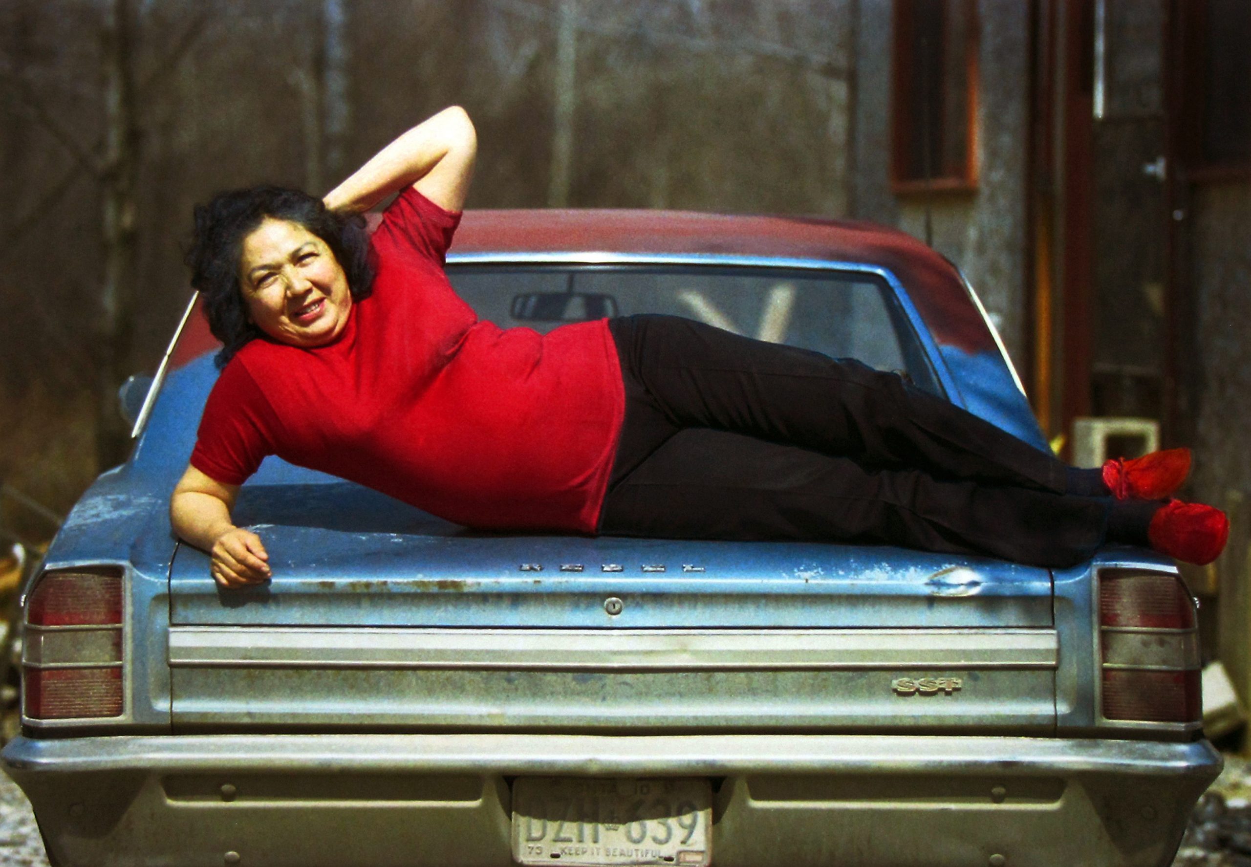 A woman in a red shirt lays sideways on the trunk of an old car, smiling.