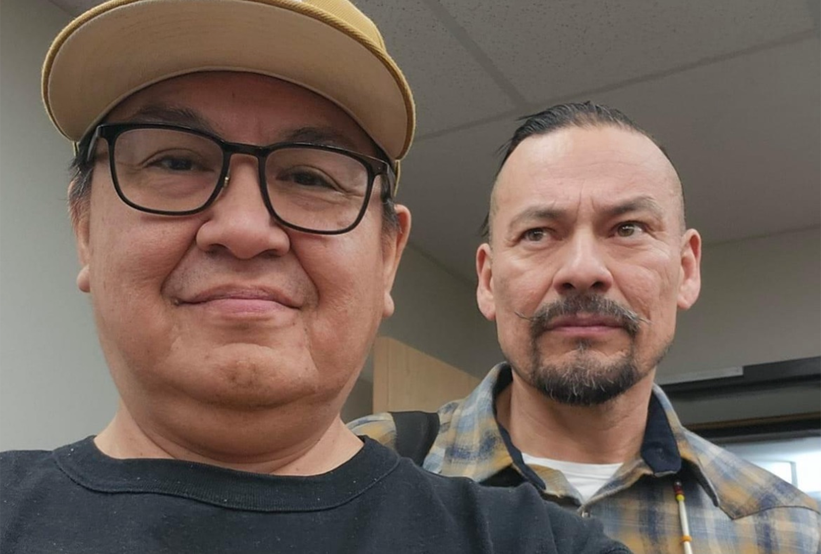 Two adult men in an indoor setting. The man in front is taking a selfie, wearing a light-colored baseball cap, black-rimmed glasses, and a black T-shirt. He’s smiling. Behind him, another man in a plaid shirt has a mustache and a short goatee, looking off to the side.