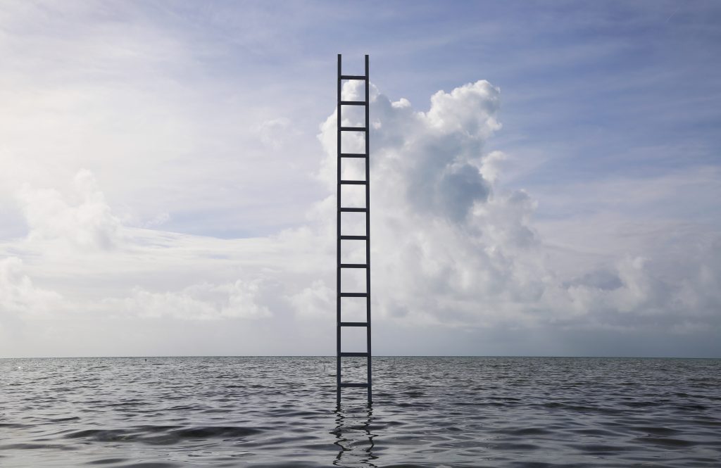 A tall, black ladder stands alone in the middle of a calm ocean, extending straight up into the sky. The water is gently rippling beneath it, and the horizon is visible in the distance. Above, soft clouds fill the blue sky, giving the impression that the ladder might lead to the heavens. The image evokes a surreal, dreamlike atmosphere.