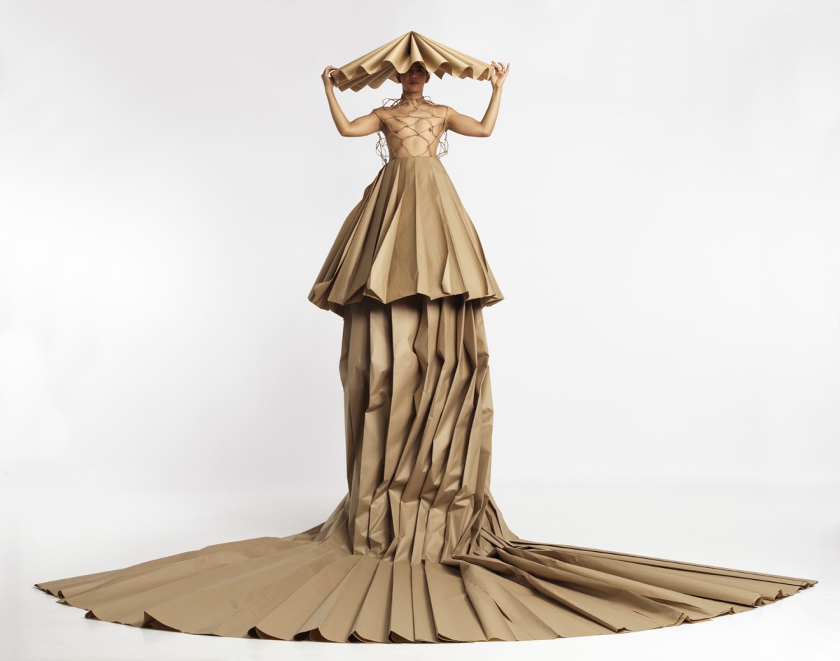 A performer stands against a plain white background wearing an elaborate, sculptural outfit resembling folded kraft paper. The towering garment features dramatic pleats that cascade to the floor in a long train. The top includes a netted bodice and a structured, fan-like headpiece held above the performer’s head. The design blends fashion, sculpture, and performance art.
