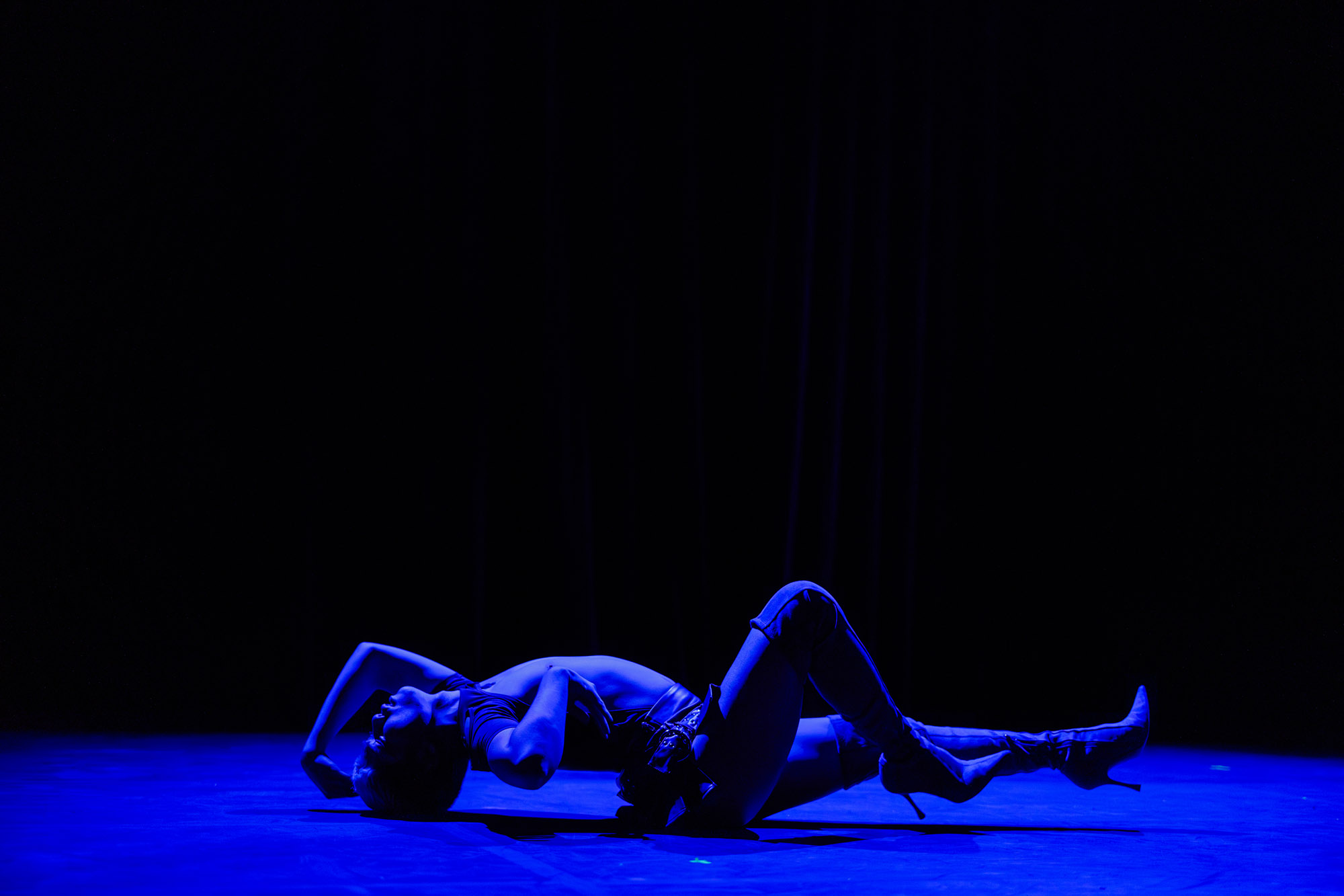 A performer lies on their back on stage under deep blue lighting, one arm raised behind their head and one leg bent, wearing high-heeled boots in a dramatic, expressive pose.