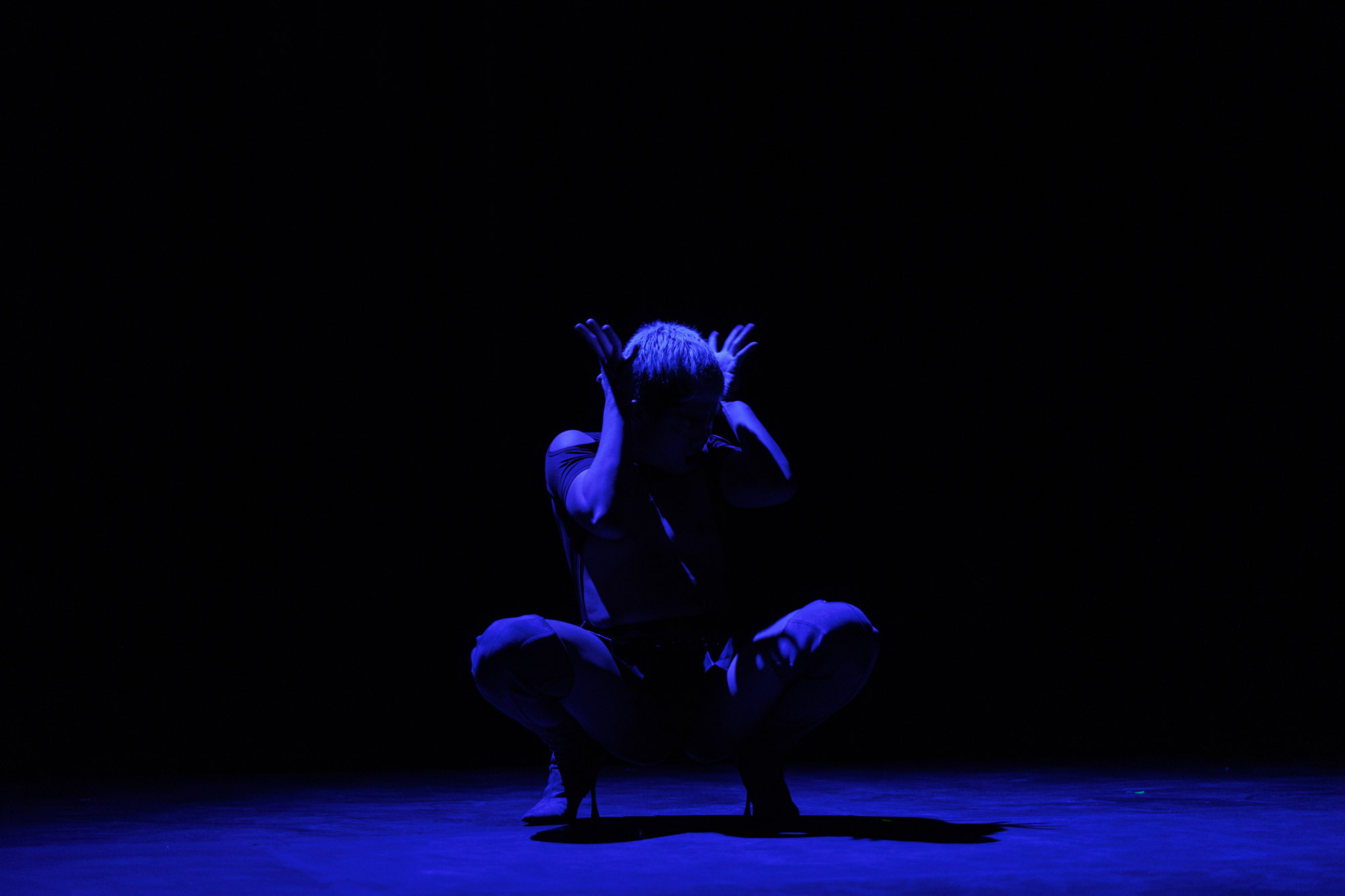 A performer crouches low on stage, dramatically lit in deep blue light against a black background, with hands raised near their head in an expressive pose.