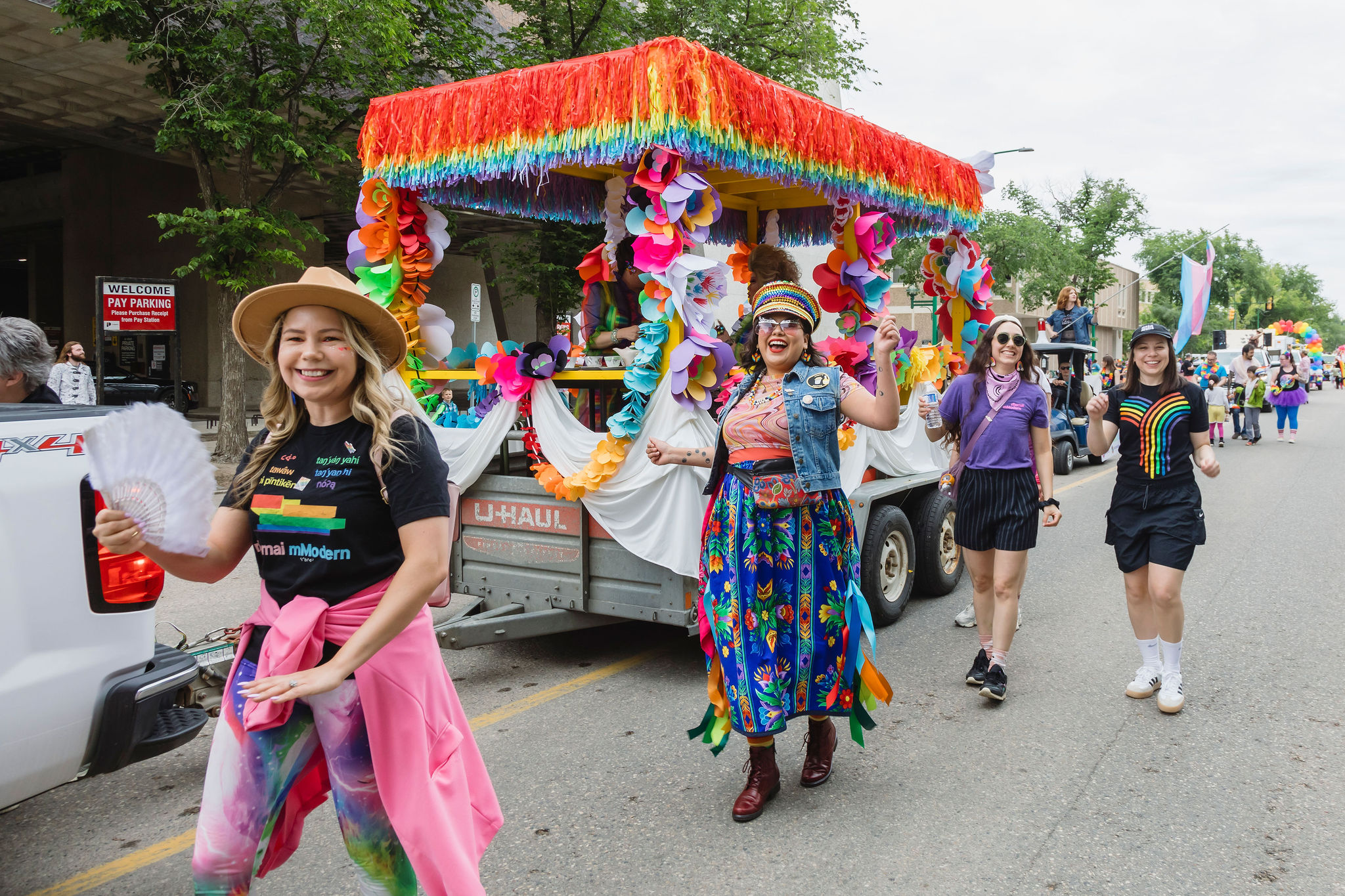 A vibrant Pride parade scene featuring smiling participants walking beside a decorated float. The float is covered in rainbow fringe and large colorful paper flowers. One person in front wears a straw hat, rainbow leggings, and a Remai Modern T-shirt, waving a white fan. Another wears a multicolored skirt and vest, dancing joyfully. Two more participants walk alongside, wearing rainbow-themed shirts and accessories. The atmosphere is festive and celebratory, with others visible in the background and a “Pay Parking” sign on a nearby building.